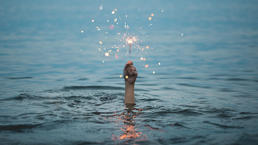A hand appearing out of a lake of water, holding a lit sparkler.