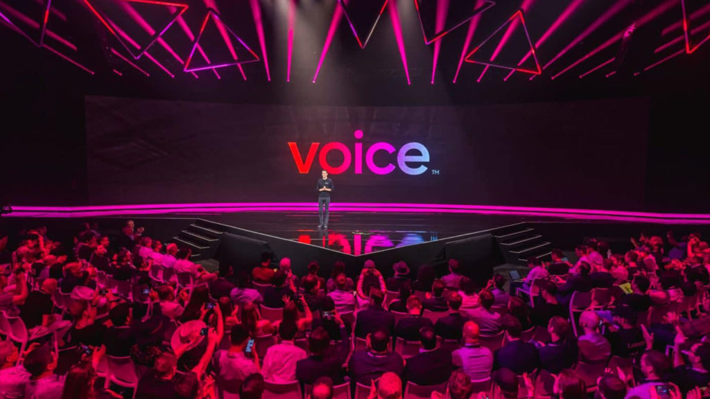 A photograph of an event for VOICE, a Blockchain-powered social network, in an auditorium filled with people, with a single male individual standing on a black, reflective stage, with the words, "VOICE" spelled out in red, pink, and blue behind him.