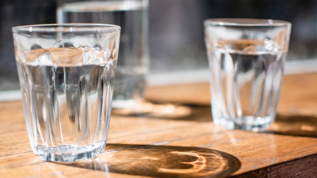 2 clear glasses of water, and a clear water pitcher in the background, all sitting on a wooden table, casting shadows and light refraction.