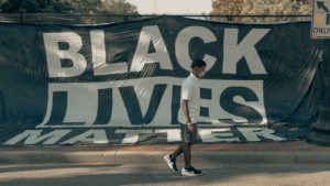 A young Black man with dreadlocks in a white button-up, short-sleeved shirt, olive green cargo shorts, and white & black nike low-tops, wearing a PPE mask, walking in front of a large "Black Lives Matter" sign draped over a black metal chain-link fence.